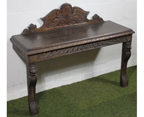 An oak console table, the raised back carved with leaves, patera, etc., with a leafy frieze on shaped legs headed by lion mas