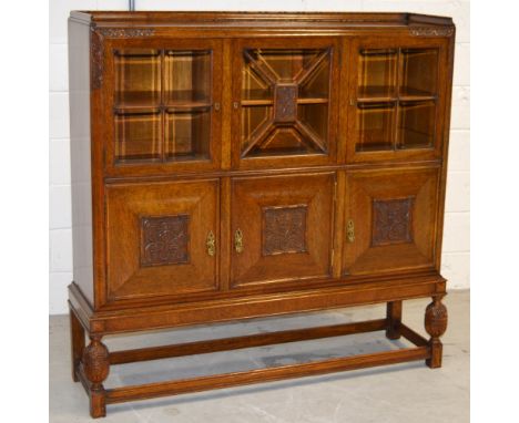 An early 20th century oak court style cupboard with three glazed doors to the top, the central door with a carved panel of vi