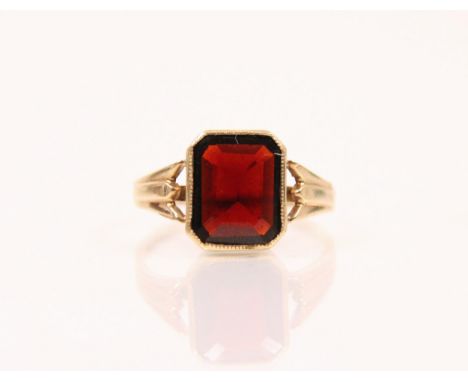 A garnet and 9ct gold ring, the emerald cut garnet within a milgrain setting edge, leading to tapered grooved shoulders and p