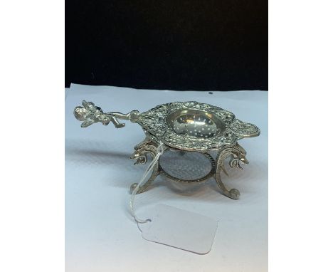 A SILVER PLATED TEA STRAINER WITH CHERUB HANDLE ON A DECORATIVE TRIPOD STAND 
