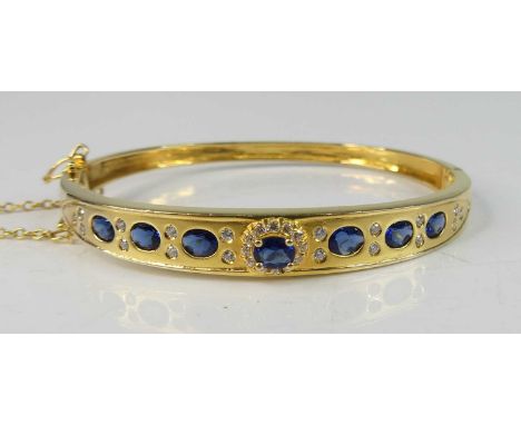 A yellow metal sapphire oval hinged hollow bangle, with box clasp, two figure-of-eight safety catches and safety chain, featu