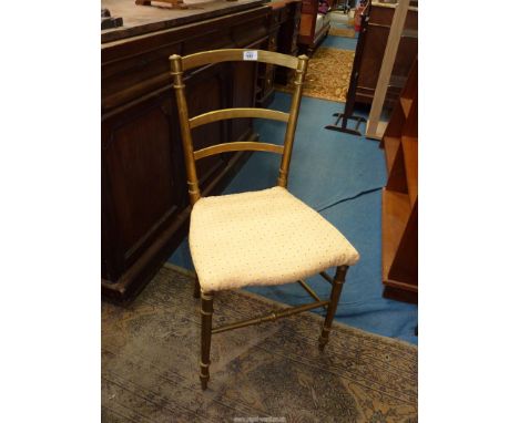 A gold painted ladder back bedroom chair standing on turned legs and having beige upholstered seat.