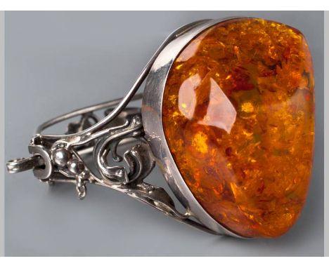 AN AMBER AND SILVER BANGLE, centre set with large organic form amber, hinged bangle decorated with scroll work, with tongue c
