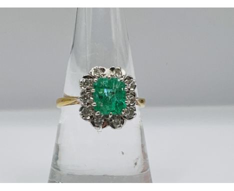  An emerald and diamond cluster ring in 18ct gold. The emerald is an octagonal step cut, measuring approximately 6.92mm x 6.2