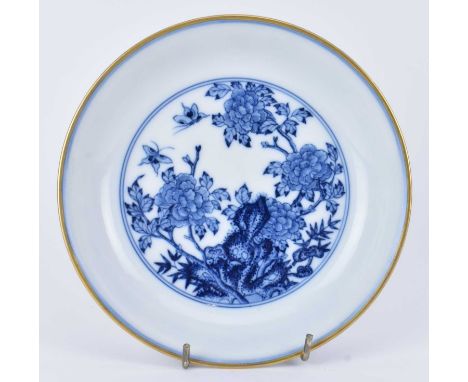 A blue and white ceramic plate with gilt rims. The circular plate decorated with peonies, butterflies, flowers and rockscape,