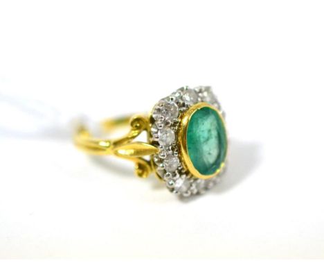 An emerald and diamond cluster ring, the oval cut emerald in a yellow rubbed over setting within a border of round brilliant 