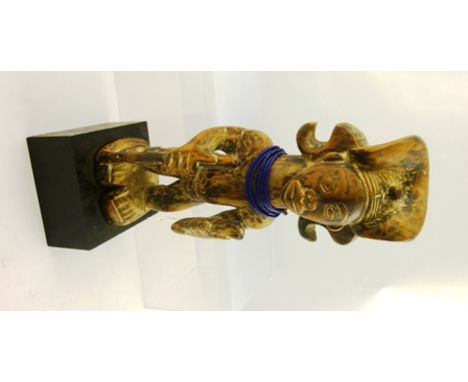 A WEST AFRICAN CARVED WOOD FETISH FIGURE carrying a stick, wearing a blue bead necklace, raised on ebonised block base, with 