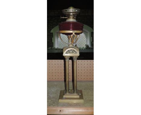 A Victorian oil lamp, the brass base raised by four reeded columns with Corinthian column caps, supporting a ruby coloured gl