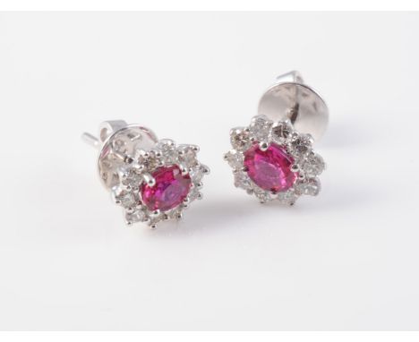 A PAIR OF RUBY AND DIAMOND EARRINGS, each oval cut ruby claw set raised within a surround of ten brilliant cut diamonds, in s