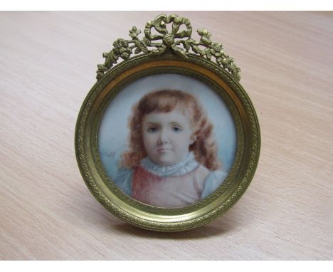A portrait on ivory of a young girl wearing a pink pinafore, decorative brass easel back frame surmounted by ribbons and flow