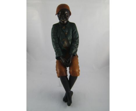 A painted figure of a black boy dressed as a jockey in tan trousers, green corduroy coat and jockey cap moulded to sit on the