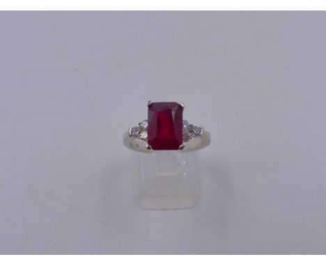 A 750 STAMPED WHITE METAL RUBY AND DIAMOND RING. THE CENTRAL RUBY IS AN EMERALD CUT IN AN ELEVATED FOUR CLAW SETTING FLANKED 