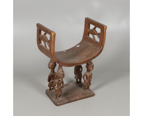 A hardwood stool with a curved seat, supported on four figures on each corner, and a rectangular base. The pierced sides with