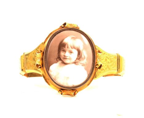 A yellow metal bangle with photograph locket centre, the oval frame to centre 40x30mm, four engraved scrolls supporting, ten 