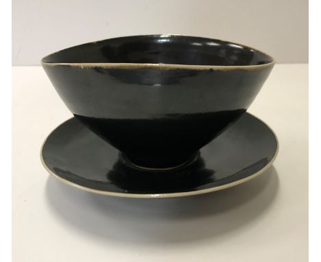 A Lucie Rie black glazed elliptical bowl and saucer, both bearing Lucie Rie marks, the bowl 14.5 cm x 12.5 cm x 7.5 cm high, 
