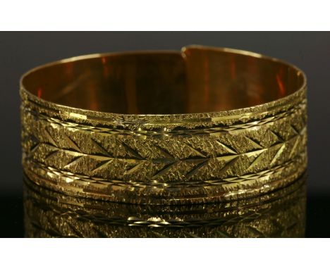 A gold, flat section sprung bangle,19.7mm wide, with diamond cut decoration to a textured ground.  Tested as approximately 18