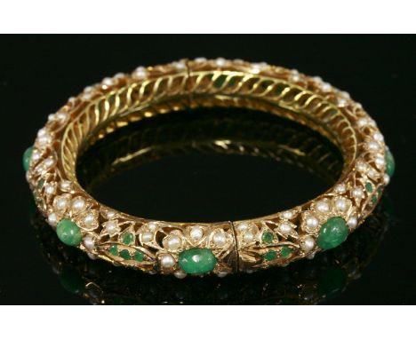 An Indian gold, emerald and pearl hinged bangle,of pierced circular section.  A series of oval cabochon emeralds, grain set t