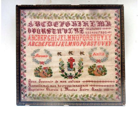 An early 20th century French needlework sampler with alphabet, numbers, cottages and crucifix within a scrolling foliate bord