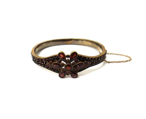 A Bohemian garnet set oval hinged bangle, the front with a flowerhead shaped motif, on a snap clasp, fitted with a safety cha