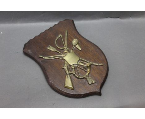 An oak plaque mounted with a brass flintlock sporting gun, a fishing net, riding crop, hunting horn, bow and arrow.  The shie