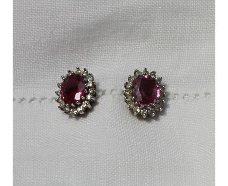 A pair of ruby and diamond cluster ear studs, each set with an oval mixed cut ruby, claw set within a surround of sixteen sma