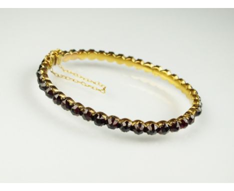 A Bohemian garnet hinged bangle, designed as a row of rose cut garnets set to yellow metal hinged bangle with attached safety