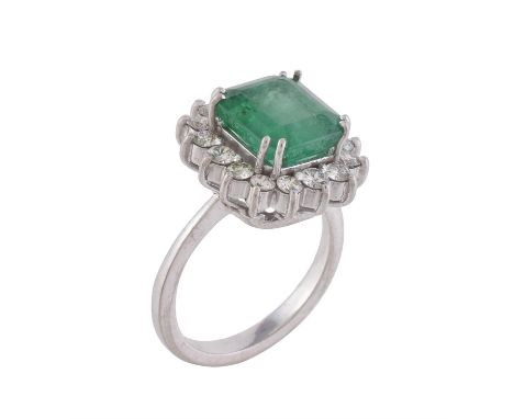 An emerald and diamond cluster ring, the step cut emerald with canted corners estimated to weigh 3.06 carats, within a surrou