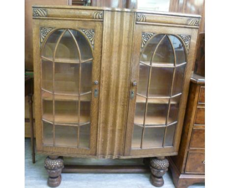 A 1930s oak bow front display cabinet with carved ornament, the pair of arched, glazed panelled doors enclosing a shelved int