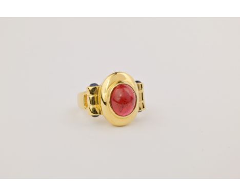 An 18ct gold, ruby and sapphire ring, featuring an oval, cabochon cut ruby held in a bezel setting atop gold scroll detailing