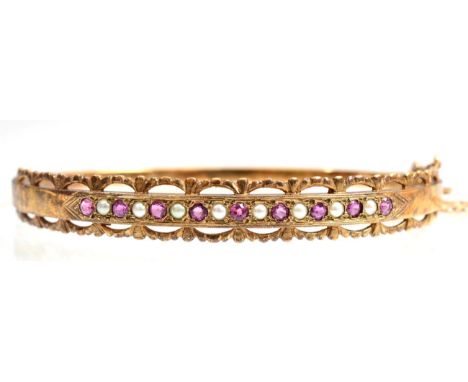 A 9 carat gold Victorian revival bangle, alternating round cut rubies and seed pearls in a scroll frame, to a plain hinged ba