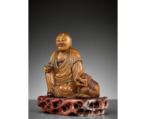 A SOAPSTONE FIGURE OF A LUOHAN WITH A BUDDHIST LION, 18TH CENTURYChina. Seated with his right knee raised, wearing long flowi