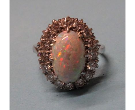 An opal and diamond cluster ring claw-set an oval opal, 12.5 x 8mm, within a surround of sixteen brilliant-cut diamonds, in 1