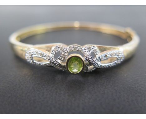 A 9 ct gold diamond and peridot hinged bracelet set with a single oval peridot - measuring approx. 6 mm by 5 mm - flanked eit