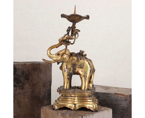 A Chinese gilt-bronze candlestick,in the form of an elephant standing on a stepped plinth, holding a stem of lotus leaves in 