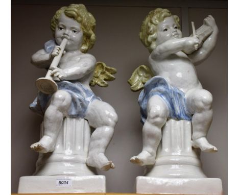 A pair of decorative ceramic cherubs, playing musical instruments, scantily clad, seated on fluted pedestals, crackle glazed,