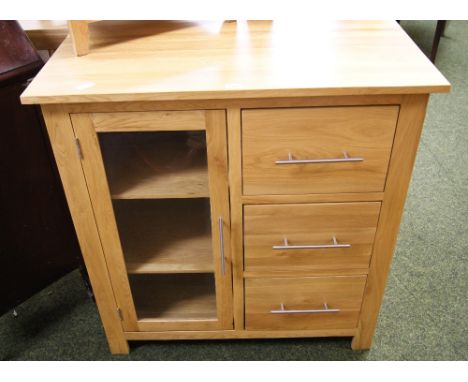 A modern, light solid oak side cabinet with a single glazed door housing shelves and three short drawers, with brushed steel 