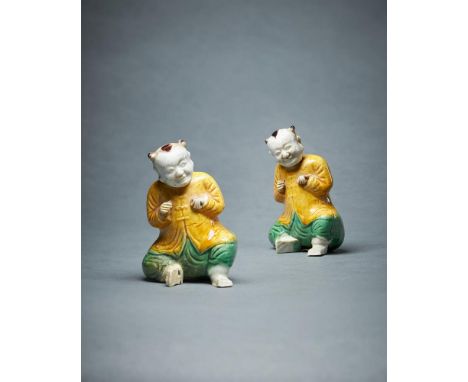 A pair of Chinese biscuit glazed models of seated smiling boys, Kangxi, with arms raised, glazed in yellow and green, each wi
