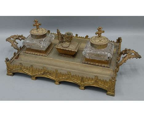 A 19th Century gilt metal inkstand with two glass inkwells and central candlestick and with shaped end handles, 41cms wide