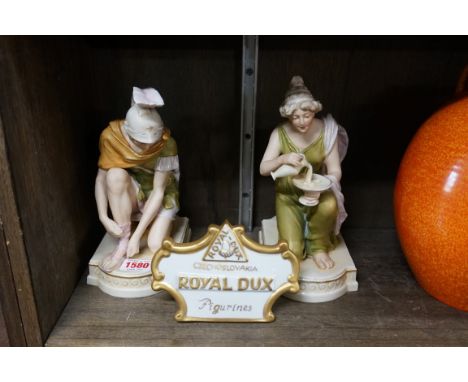 A pair of Royal Dux shot enamel figures,&nbsp;21cm high; together with a Royal Dux porcelain sign, 14cm wide.&nbsp; 