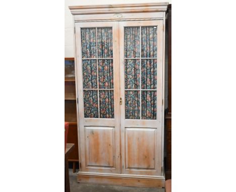A limewashed pine wardrobe with part glazed and panelled doors enclosing shelf and hanging rail fitted with floral curtains, 