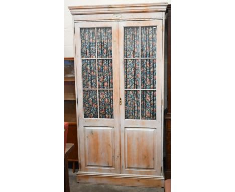 A limewashed pine wardrobe with part glazed and panelled doors enclosing shelf and hanging rail fitted with floral curtains, 