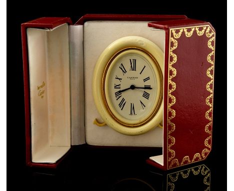 A Cartier alarm clock with circular white enamel dial, cream and gilt oval case with easel back, cabochon blue stone winders,