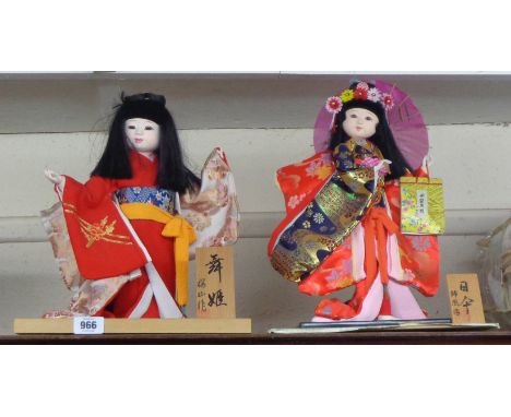 Two vintage Japanese geisha display dolls with original fabric clothing on titled wooden stands