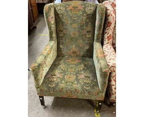 An early 20th Century box framed wing-back armchair upholstered in floral tapestry, set on square legs and casters