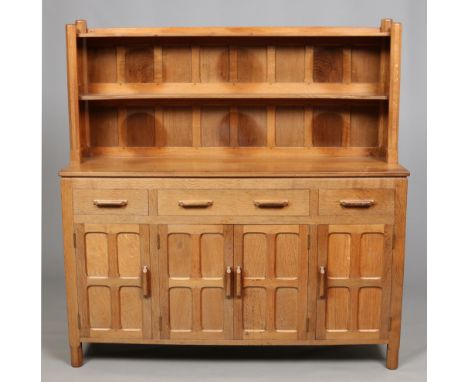 A Cotswold School light oak sideboard. With panelled delft rack and cupboard doors, having three drawers and raised on chamfe