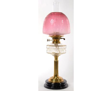 AN EDWARDIAN BRASS FLUTED COLUMNAR OIL LAMP WITH GLASS FOUNT, BRASS BURNER AND BLACK GLAZED POTTERY FOOT, ETCHED CRANBERRY GL