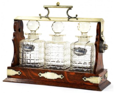 AN EPNS MOUNTED OAK TANTALUS 38CM W CIRCA 1910 WITH A SET OF THREE CUT GLASS SPIRIT DECANTERS AND STOPPERS AND A PAIR OF SILV