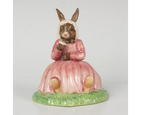 A figure of a bunny wearing a pink dress and white bonnet, sitting on the grass and holding a flower. Bunnykins of the Year 2