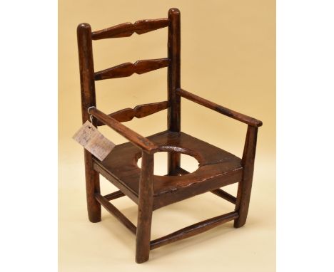 A  INFANT'S PRIMITIVE POTTY CHAIR possibly in elm, with three rung ladder back and arm supports, 33cms wide