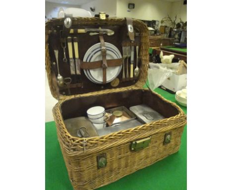 A wicker picnic hamper with leather and brass mounts opening to reveal cotents including various cutlery, enamel plates, tins